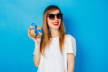 A young girl with a smile in glasses and a white T-shirt is holding a gift box on a blue background. Birthday or holiday gift concept.