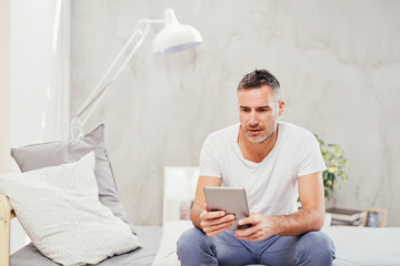 Caucasian man in forties sitting on bed in bedroom and using tablet.