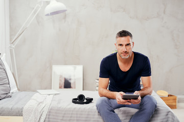 Caucasian man in forties sitting on bed in bedroom and using tablet.