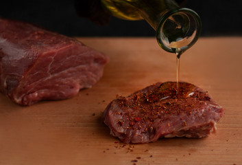 Hand in black glove pouring oil on beef steak with spices on a wooden board with a black background