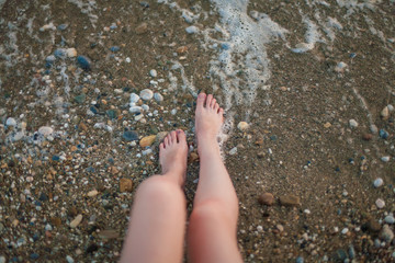 Vacation on tropical beach. Woman's legs in the clear ocean water. First person perspective. Summer holidays, Vacation concept. Girl sitting on sandy beach in water. Bare feet
