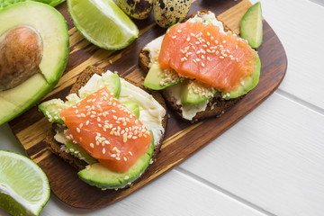 sandwiches with mashed avocado and sesame and salted salmon and half of the avocado fruit lying next to it on a wooden board on a light wooden background close-up