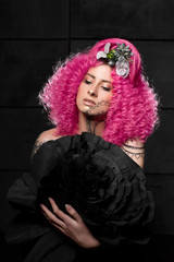 young attractive caucasian girl model with afro style curly bright pink hair, tattooed face and flowers woven into her hair. Photo in the studio on a black background