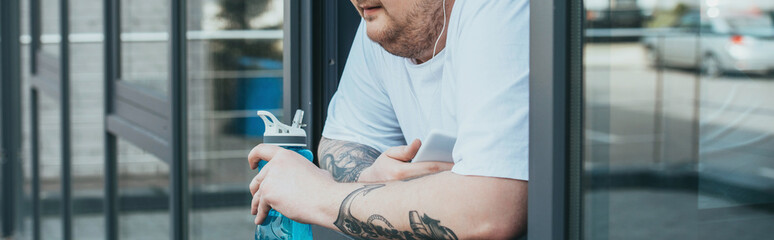 Fototapeta premium panoramic shot of overweight man with earphones and smartphone holding sport bottle and looking out through window at gym