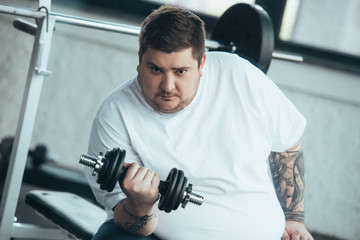 Fototapeta na wymiar Overweight tattooed man Looking At Camera and training with dumbbell at sports center
