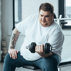 Fototapeta na wymiar Overweight tattooed man sitting and exercising with dumbbell at sports center
