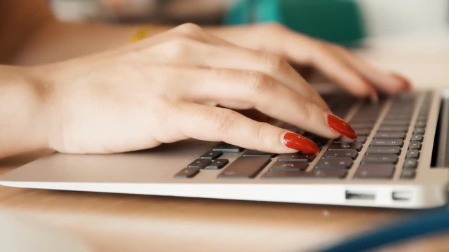 Handheld slow motion shot: the hands of a woman typing on a laptop's keyboard. Long manicured nails with red polish.