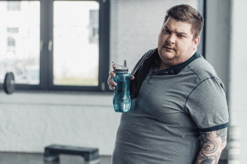 overweight tattooed man looking at camera and holding sport bottle at gym