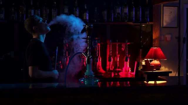 Confident man resting in lounge restaurant on against black background, smoking hookah. A young man smokes a hookah in a hookah bar on the background of a bar with hookahs in the dark, slow motion.