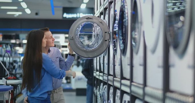 A handsome man and a woman choose a washing machine to buy open the door and inspect the coating of the drum and the quality of the product. Woman looking inside washing machine