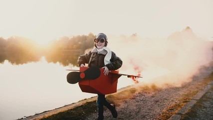 Happy little pilot girl playing, running along sunset lake in fun cardboard plane costume with color smoke slow motion. - Powered by Adobe