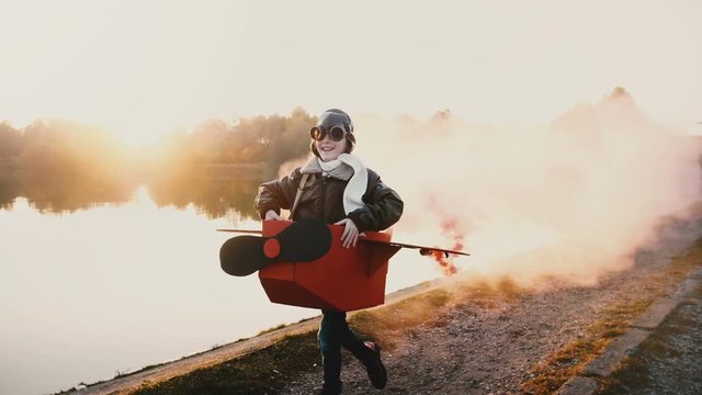 Happy little pilot girl playing, running along sunset lake in fun cardboard plane costume with color smoke slow motion.