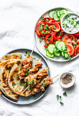 Greek chicken skewers, flatbread, tomatoes, cucumber salad, baked sweet pepper, tzatziki yogurt herb sauce on a light background, top view. Mediterranean style lunch table