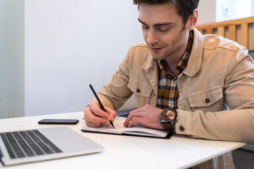 Concentrated freelancer in jacket writing in notebook at table