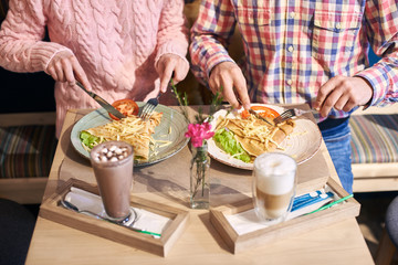 Cropped view of young couple, friends sitting together in cafe at small table enjoying big pancake sandwich with vegetables. Coffee beverage and vase with flower on blurred foreground. High angle view