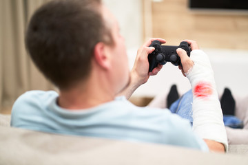 Man with broken arm in plaster cast with red blood play and holding controller in front of TV. Game addiction concept.