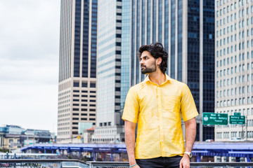 Raining day - grainy, wet feel. Young East Indian American Man with beard traveling in New York City, wearing yellow short sleeve suit, standing in front of modern high buildings, looking around..