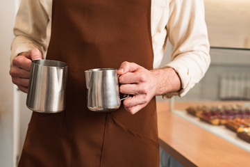 Cropped view of barista in brown apron holding steel milk jugs