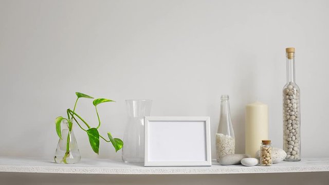 Modern room decoration with picture frame mockup. Shelf against white wall with decorative candle, glass and rocks. Hand putting down plant cuttings in glass.