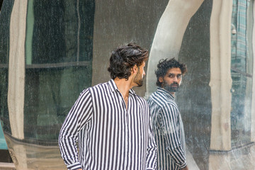 Obraz na płótnie Canvas Raining day - grainy, drizzling, wet feel. Young East Indian American Man in New York, with beard, wearing black, white striped shirt, standing outside, turning around, looking reflection on mirror..
