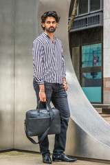 Raining day - grainy, drizzling, wet feel. Young East Indian American Man with beard, wearing black, white striped shirt, black pants, leather shoes, holding bag, standing on street in New York..