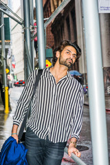 Raining day - grainy, drizzling, wet feel. Young East Indian American Man with beard, wearing black, white striped shirt, carrying shoulder bag, holding blue clothes, standing on street in New York..