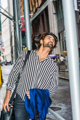 Raining day - grainy, drizzling, wet feel. Young East Indian American Man with beard, wearing black, white striped shirt, carrying leather shoulder bag, carrying blue clothes, looking up in New York..