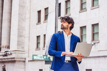 Way to Success. Young East Indian American Businessman with beard traveling, working in New York, wearing blue suit, holding laptop computer, standing on Wall Street, confidently looking up..