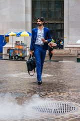 Raining day - grainy, foggy, wet feel. Young East Indian American Business Man with beard, wearing blue suit, white shirt, carrying leather hand bag, holding coffee cup, walking on street in New York