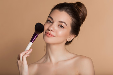 Closeup portrait of beautiful happy young girl with perfect skin is holding makeup brush in hand. Isolated on beige. Beauty and cosmetics concept.