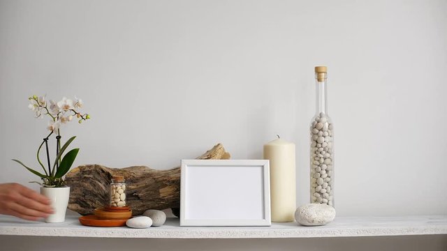 Modern room decoration with picture frame mockup. Shelf against white wall with decorative candle, glass and rocks. Hand putting down potted orchid plant.