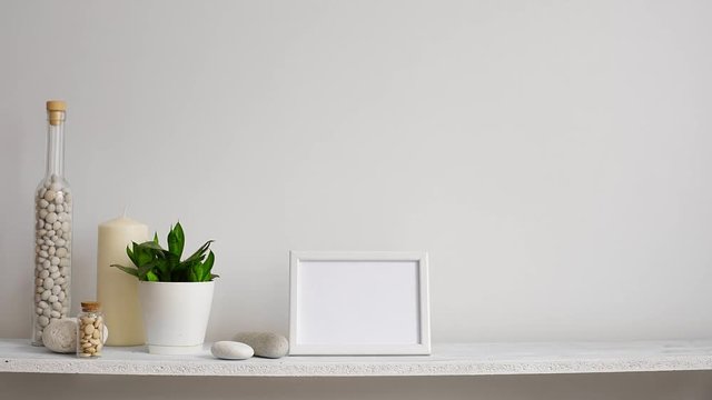 Modern room decoration with picture frame mockup. Shelf against white wall with decorative candle, glass and rocks. Hand putting down potted snake plant.