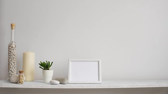 Modern room decoration with picture frame mockup. Shelf against white wall with decorative candle, glass and rocks. Hand putting down potted succulent plant.