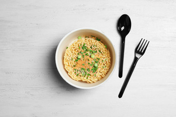 Cup of instant noodles with plastic cutlery on white wooden background, flat lay