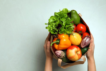 Woman holding paper bag with fresh vegetables and fruits on light background, top view. Space for...