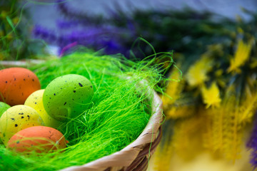 Fototapeta na wymiar Green nest in a basket with small colorful Easter eggs, decoration, close-up easter concept, holiday tradition, blurred paints and plastic flowers on yellow background, selective focus