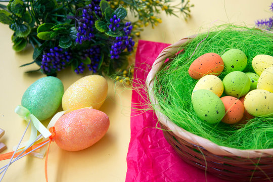 Green nest in a basket with small colorful Easter eggs, decoration, close-up easter concept, holiday tradition, blurred paints and plastic flowers on yellow background, selective focus