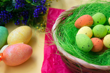 Green nest in a basket with small colorful Easter eggs, decoration, close-up easter concept, holiday tradition, blurred paints and plastic flowers on yellow background, selective focus