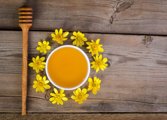 honey in the glass bowl, dipper and yellow flowers around it on wooden background