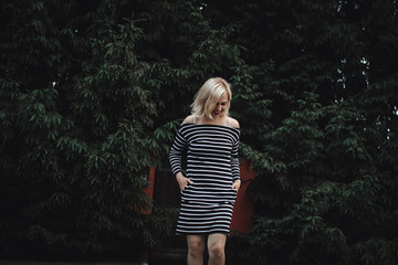 Stylish interesting blonde girl in a striped dress at a wooden fence and a thick hedge of fir trees. Fun and cute smiles.