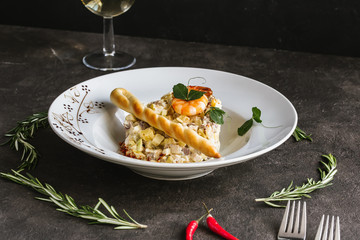 Traditional Russian salad Olivier on a white plate with breadsticks and shrimp with white wine glass and pepper on dark table