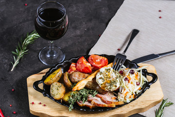 Hot assorted meats, bacon, meat sausages with roasted tomatoes, potatoes, poached egg and fresh cabbage on a platter, with a glass of red wine on a dark background