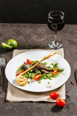 Traditional German fried pork cheeks with greens and tomato closeup on a white plate with bread cakes on a served table with a glass of red wine