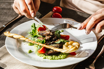 Female hands holding a fork and a knife above, eating traditional German fried pork cheeks with greens and tomato close-up on a white plate with bread cakes on a served table with a glass of red wine.