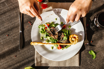 Female hands holding a fork and a knife above, eating traditional German fried pork cheeks with greens and tomato close-up on a white plate with bread cakes on a served table with a glass of red wine.