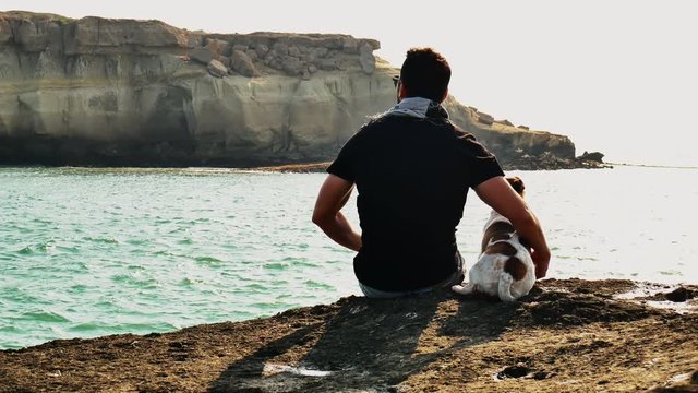 A panning shot of from back of a man and his dog sitiing on edge of rock  A shot of a young man and his jack russel dog sitting on edge of rock on persian gulf ocean sunny natural light