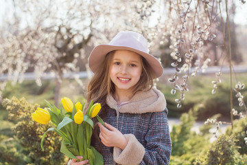  Beautiful girl smiling. A girl with long hair in a pink hat and coat stands on the street. The child is holding yellow tulips