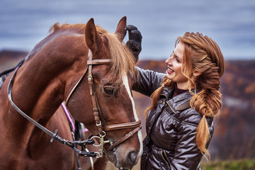 Obraz premium A young woman with brown curly hair caresses a brown horse. The horse caresses the woman. Woman on a walk with a horse on a cloudy autumn day.