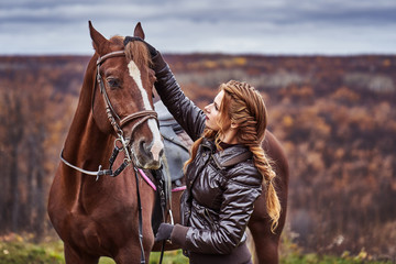 A young woman with brown curly hair caresses a brown horse. The horse caresses the woman. Woman on...