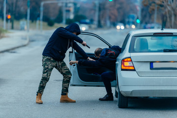 The car thief is pulling the car owner out of his car and trying to get the car while pointing a...